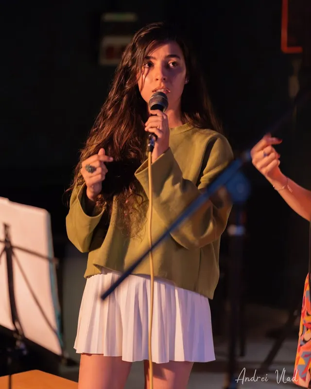 Olivia Mihet cântând la microfon, într-un pulover verde, sub o lumină caldă de scenă