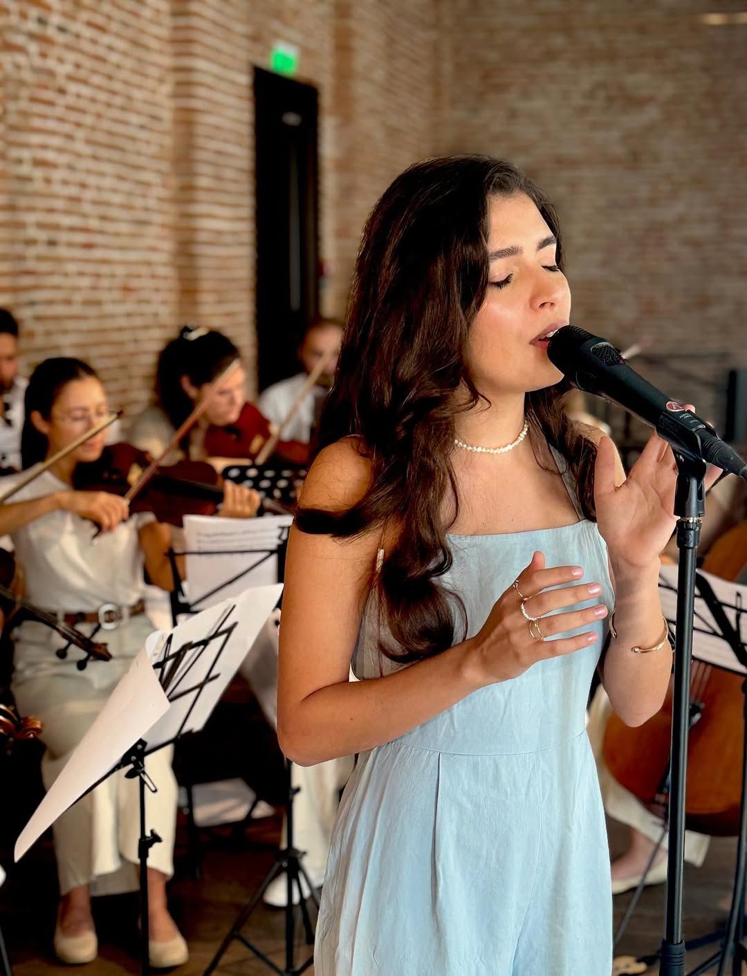 Portret apropiat al Oliviei Mihet cântând cu ochii închiși, cu violoniști vizibili în spatele ei, la același concert