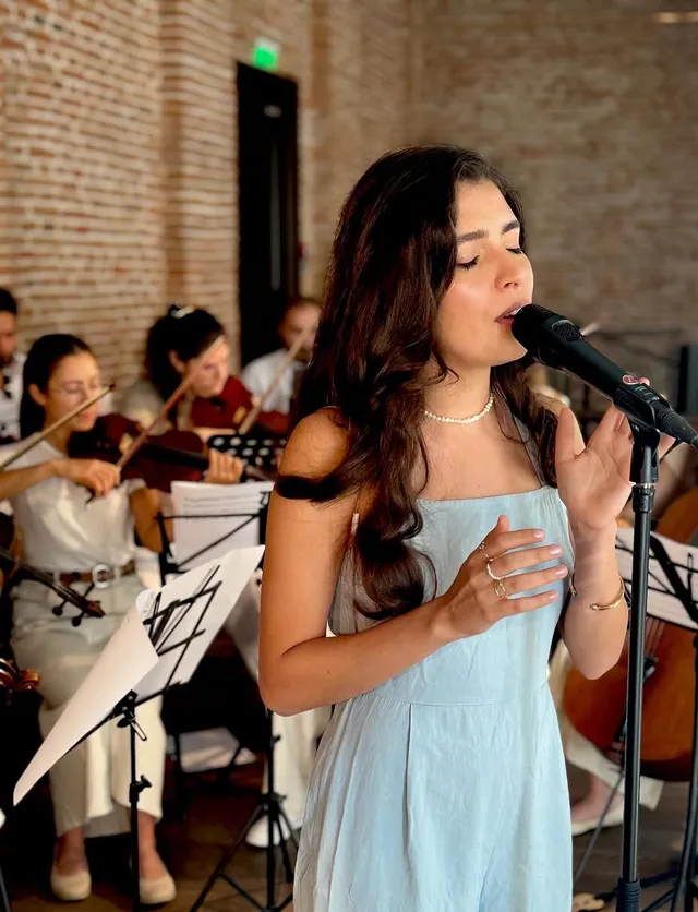 Portret apropiat al Oliviei Mihet cântând cu ochii închiși, cu violoniști vizibili în spatele ei, la același concert