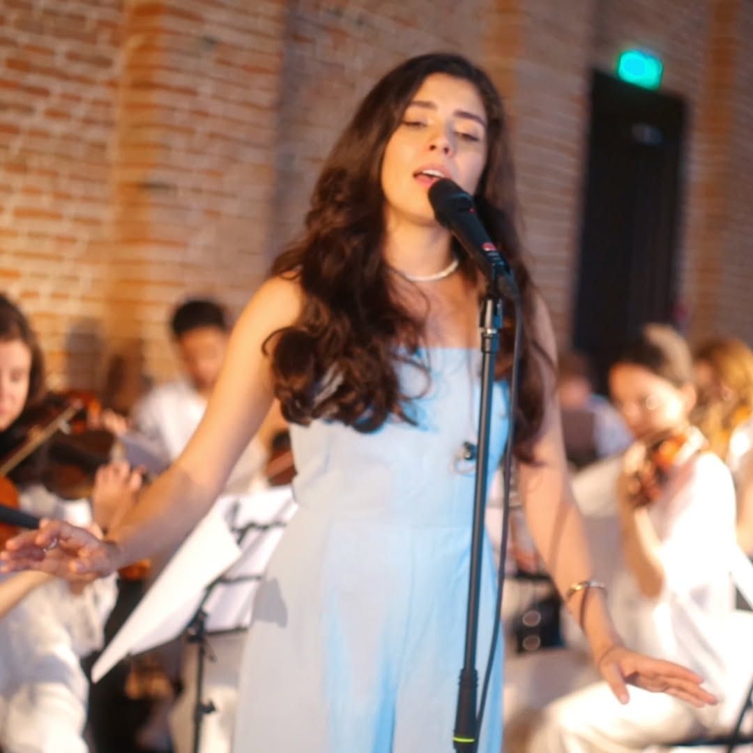 Olivia Mihet singing with arms open on stage with a string ensemble in a brick-walled hall