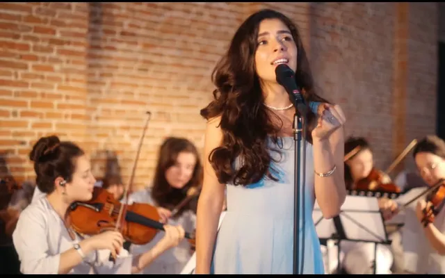 Olivia Mihet într-o rochie bleu pal, cântând alături de un ansamblu de coarde într-o sală de concert din cărămidă aparentă, luminată cald