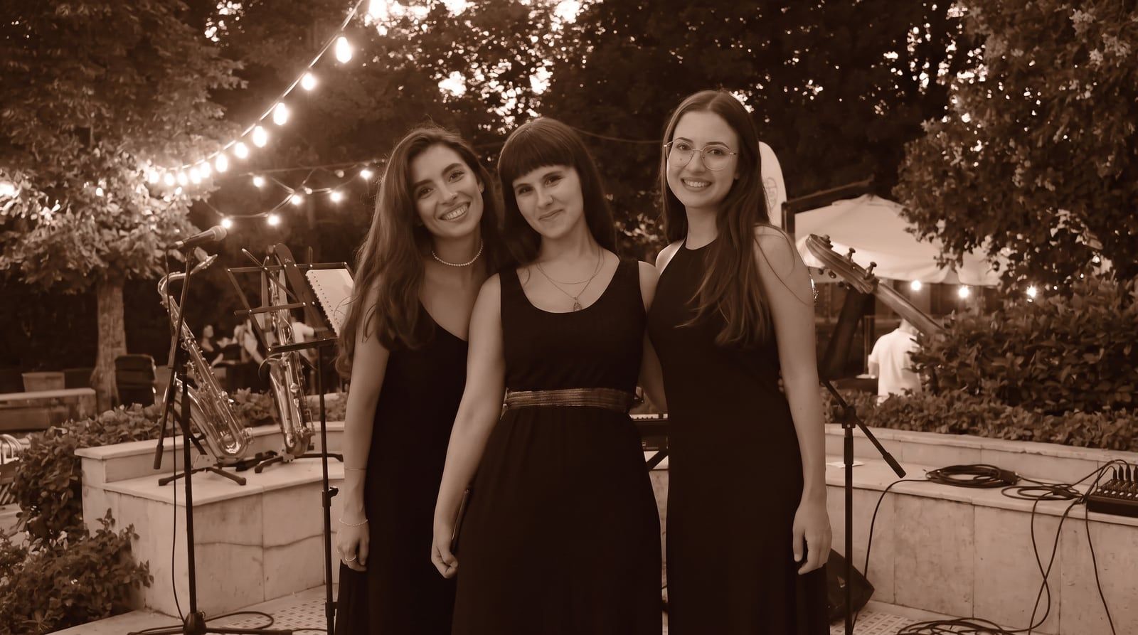Olivia Mihet cântând într-un trio, în rochii negre, pe o scenă de grădină cu ghirlande luminoase, cu un saxofon pe suport alături