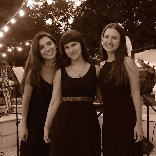 Olivia Mihet cântând într-un trio, în rochii negre, pe o scenă de grădină cu ghirlande luminoase, cu un saxofon pe suport alături
