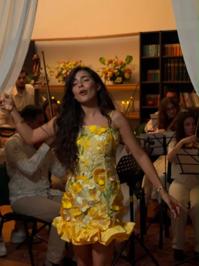 Olivia Mihet într-o rochie galbenă cu model floral, cântând într-o încăpere tip bibliotecă, cu coarde, draperii transparente și lumânări
